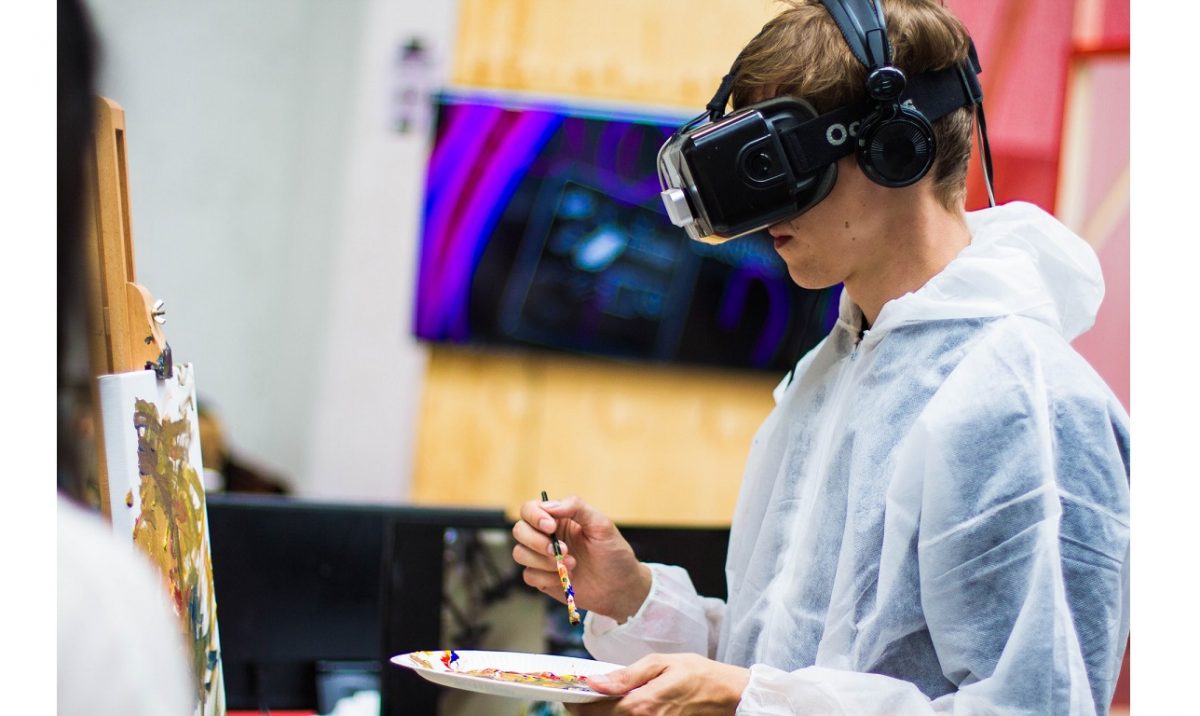 Kuvituskuva. Henkilö seisoo taulucanvaksen edessä maalipaletti ja sivellin kädessä, VR-lasit päässään.